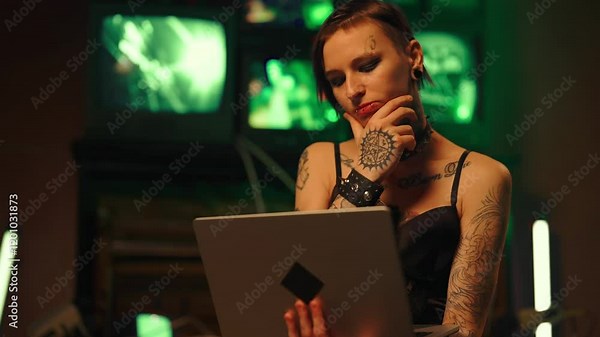 Tattooed woman hacker working with laptop in basement in night, cybercrime. Alternative style lady with piercing and tattoo surfing internet by portable computer, hacking and programming web code