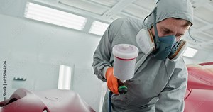 Auto body technician in protective gear spray painting a red car bumper in a specialized paint chamber at a car service center. Car Painter in Protective Gear Spray Painting Red Car Bumper