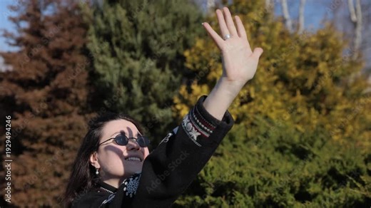Happy young woman in sunglasses rises hand with splayed fingers to cast shadow on face in spring park. Female in embroidered jacket smiles to sun standing among green garden. Springtime vibes