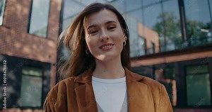 Camera zooming in on gorgeous Caucasian girl. Woman looking at camera and smiling widely. Wearing golden earrings and leather jacket on top of white shirt. Posing for best shot in front of building.