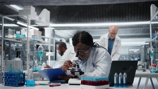 Medical laboratory expert using scientific microscope for cellular analysis, writing down research findings. African american woman examining sample in lab with optical device gear, camera A
