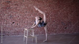 Pretty female child artistic gymnast in performance suit make warm-up in studio and perform muscle stretching exercise, leaning on stool. Rhythmic gymnastics for girls