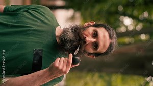 vertical video portrait of a bearded adult man holding a pipe with tobacco in a city park exhaling smoke smoking bad habits unhealthy lifestyle