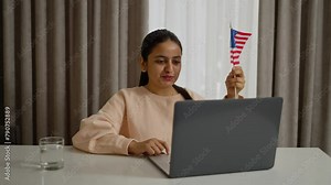 Happy brunette girl with Brown skin color studies foreign languages at home using her laptop and communicates via video conference while holding the flag of the United States of America at home in a