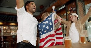 Happy mixed race supporters cheering and exulting after winning. Friends at the bar in a pub having fun. Men with the flag of the national country of USA drinking beer and watching sport game.