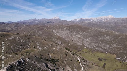 Ultra-wide aerial panorama from Rocca Calascio area showing vast Abruzzo mountain landscape with rolling valleys, winding roads, and majestic snow capped Gran Sasso peaks.