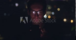 Portrait of young man using mobile phone, scrolling app pages social networks, mobile screen reflected on his face. Male in glasses scrolling internet on mobile phone background of night city.