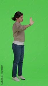 Vertical video: Side view of woman doing stop talking sign with palm over green screen, full body backdrop of model posing with rejection and denial gesture. Person raising arm to express refusal