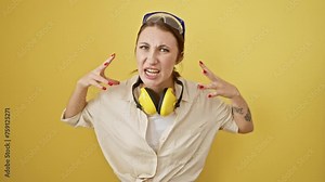Flustered young brunette girl, distressed and yelling, lets out a mad shout wearing safety glasses and noise reduction headphones, arms flailing in sheer frustration. isolated on a yellow background.