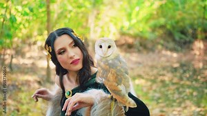forest nymph fantasy woman with white bird, holding owl in hands sits on ground fallen leaves. Green warm coat medieval style. Forest autumn trees nature river sun light. Long black hair golden diadem