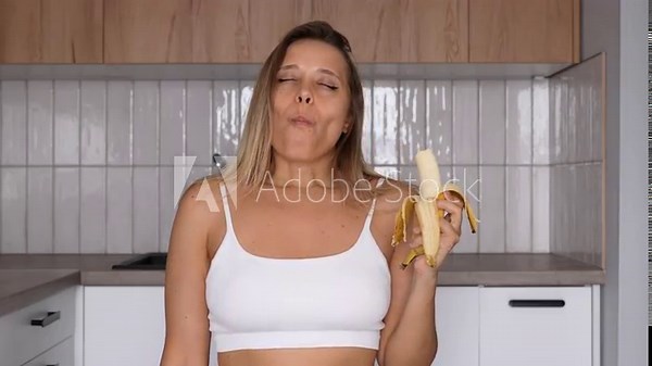 A young blonde woman in the kitchen opens a banana and eats it with visible pleasure on her face. A sensual and joyful moment of enjoying simple things in everyday life