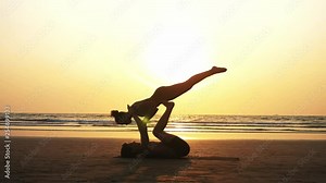 Silhouette of fit sporty couple practicing acrobatic yoga with partner together on the beach in slow motion.