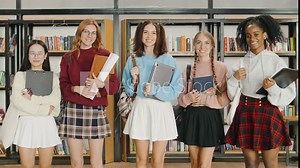 A portrait of five happy multiethnic female students standing in a university library, holding backpacks, a poster board, and textbooks, all smiling and looking at the camera.