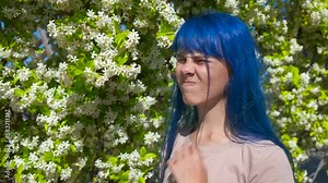 Skin itching. A young girl with blue hair scratches her itchy neck against the backdrop of a flowering plant in a spring park. Pollen allergy concept.