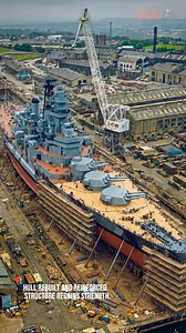 The USS Iowa (BB-61), the lead ship of the Iowa-class battleships, is shown in dry dock at a Navy shipyard undergoing maintenance and inspection. With the water drained, the ship’s massive hull rests on keel blocks, revealing features normally hidden below the surface—such as the bow shape, armor belt, propeller shafts, and rudders. Scaffolding, cranes, and work platforms surround the vessel as shipyard crews perform structural repairs, repainting, and system overhauls. The scene highlights the 