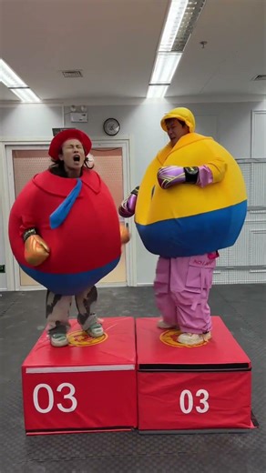 Two friends wear giant inflatable suits to battle each other on red platforms.