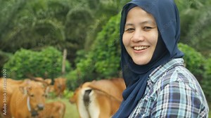 Happy successful Asian muslim female farmer looking at camera and smiling, with domesticated cow ox cattle grazing in the background, agriculture farming entrepreneur