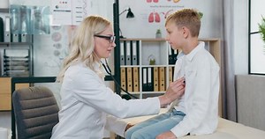Doctor visit concept where good-looking positive teen boy sitting on reviewing table when pretty smiling skilled blond female doctor checking his lungs breath and after inspection giving him high five