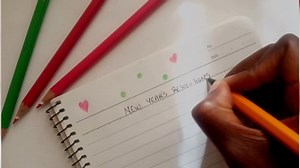 Flat lay top view of a female Africa hand writing new years resolutions words with a sharp pointed pen on a notebook and there are colored pencils near it. Dots and love hearts drawn on the notebook.