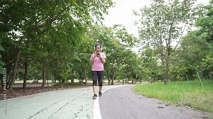 A front view of a young adult woman walking for exercise on a park road, using her smartphone, wearing athletic attire and headphones, and embracing a healthy lifestyle