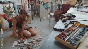 Young barefoot female artist drawing picture on paper with crayons while sitting on the floor in studio