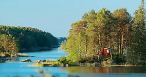 Sweden. Amazing Red Swedish Wooden Log Cabin House On Rocky Island Coast In Summer Sunny Evening. Lake Or River Landscape. Bold Colors.