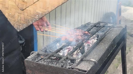 Man grilling sate maranggi on a charcoal grill in West Java, Indonesia, with a rustic background and smoke rising.