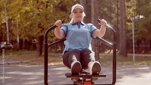 Elderly senior woman goes in for sports on a simulator doing rowing exercise outdoors in the park. Wellness procedures in retirement age. Health care and healthy lifestyle concept.