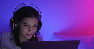 Woman gamer in bed. Caucasian woman on bed playing multiplayer game on laptop at night with color contrast. Happy talking. Front shot with copy space.