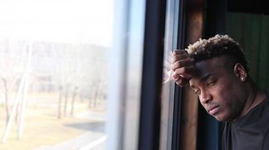 A brooding African guy in a black T-shirt is standing by the window. A black man is immersed in his thoughts, thinking about a goal, a future project, a desire for a career.