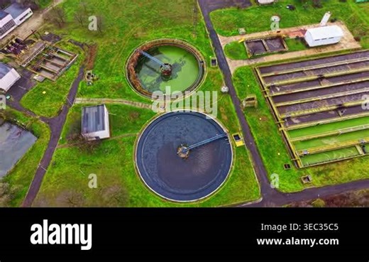 A wastewater treatment plant with round and rectangular tanks. The area is green with buildings and machinery scattered around. The image is taken from above Stock Video Footage - Alamy