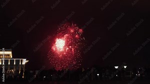 Handheld shot of real fireworks at the city fire festival, wide shot. People watch bright pyrotechnic show on the site near illuminated building. Volleys explode in the black night sky.