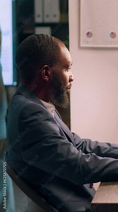 Vertical video Experienced computer operator sitting at desk during nightshift, working on company accounting payroll reports and tables in modern office while coworker in background comes back from