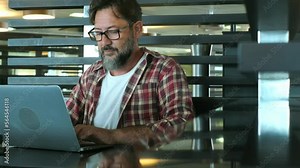 Focused young man wearing glasses using laptop, typing on keyboard, writing email or message, chatting, shopping, successful freelancer working online on computer, sitting in modern office desk