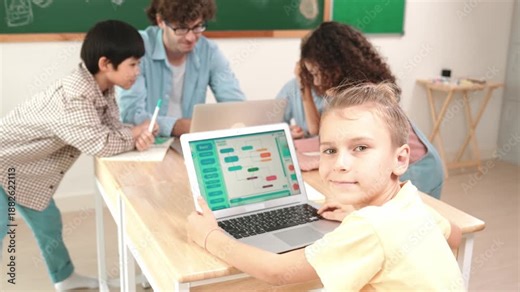 Cute caucasian boy coding program and looking at camera in STEM technology class while group of diverse student and teacher doing mind mapping. Smart child learning coding software program. Pedagogy.