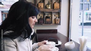 Young woman looking outside through the window in a cafe. Beautiful brunette drinks coffee from a cup, sitting indoor in trendy urban cafe.