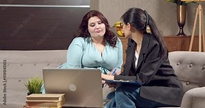 Two young women are discussing something while sitting on the couch. Attractive plus size girl talking to female psychologist or nutritionists in office