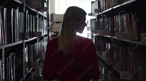 Young student walks through library choosing a book, rear view. Blonde student walks down the corridor of the library, looking around