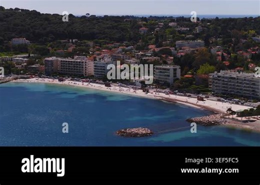 Aerial panoramic sweep of Antibes crescent beach and marina, turquoise water, rocky breakwaters, circular stone islet, hotels, apartments, pine hills, yachts, sunbathers Stock Video Footage - Alamy