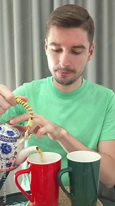 Vertical shot of Caucasian Man pouring tea from a ceramic teapot into a red mug beside a green mug on a glass table. Indoor tea preparation and hospitality concept
