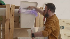 Middle-aged man wearing casual clothes coming to wooden shelves with transparent trash container in his hands and starting dropping different garbage in special containers