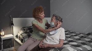 Female nurse assistant worker putting face mask on senior woman at home. Mature woman in protective mask helps put on mask to elderly female. Old mother and older daughter wearing medical masks.
