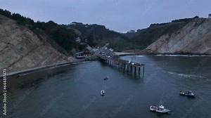 Aerial northern California Point Arena fishing pier sunset. Bay surrounded by the Pacific Ocean and coastal mountains. Recreation and commercial fishing pier and marina. Historic marine buildings.