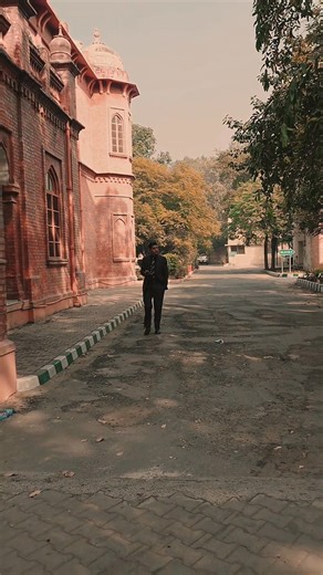 The video is a visual tour of the Chamba House (Federal Lodge) in Lahore. It primarily features the magnificent red-brick architecture of the historic building, showcasing its grand central archway entrance and a long facade lined with smaller arches and columns. The setting is bright and serene, featuring well-maintained green lawns, trees, and paved walkways. A person in a dark suit is seen walking along the path, highlighting the scale of the heritage structure. It captures the classic Indo-M