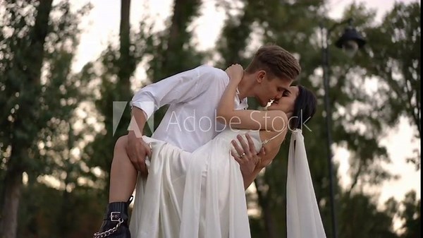 Bride in flowing white dress and groom in casual shirt kiss passionately as he holds her in his arms. Captured in slow motion, the scene highlights romance, passion, and intimacy.