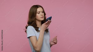 Attractive woman in blue T-shirt making voice records on mobile phone, talking to smart device, using virtual assistant for online search. Indoor studio shot isolated over pink background.