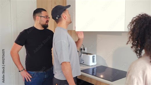 Handyman with toolbox talks to homeowners about kitchen cabinet alignment and installation process during renovation consultation inside contemporary apartment interior.
