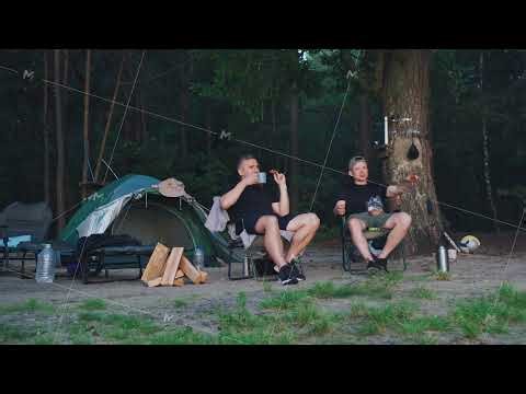 Two caucasian men camping by tent sipping coffee and sharing snacks on folding chairs beside stacked