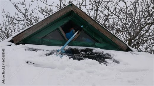 A girl cleans a solar panel from a large amount of snow on the roof of a house using a mop through a small viewing window