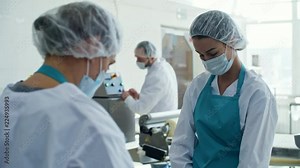 Three colleagues in protective clothing, aprons, bouffant mob caps, and face masks working in food production line at factory
